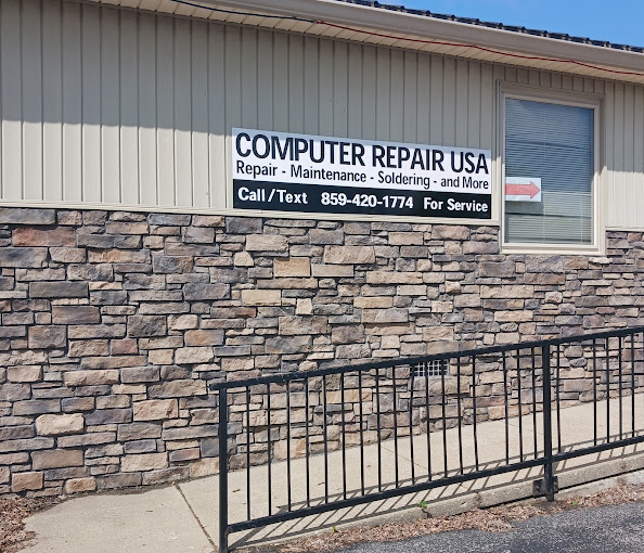 A photo of Computer Repair USA's sign attached to a building and walkway, the building is half tan and brick, the walkway is a slopped handicapped walkway, with a black guardrail.
